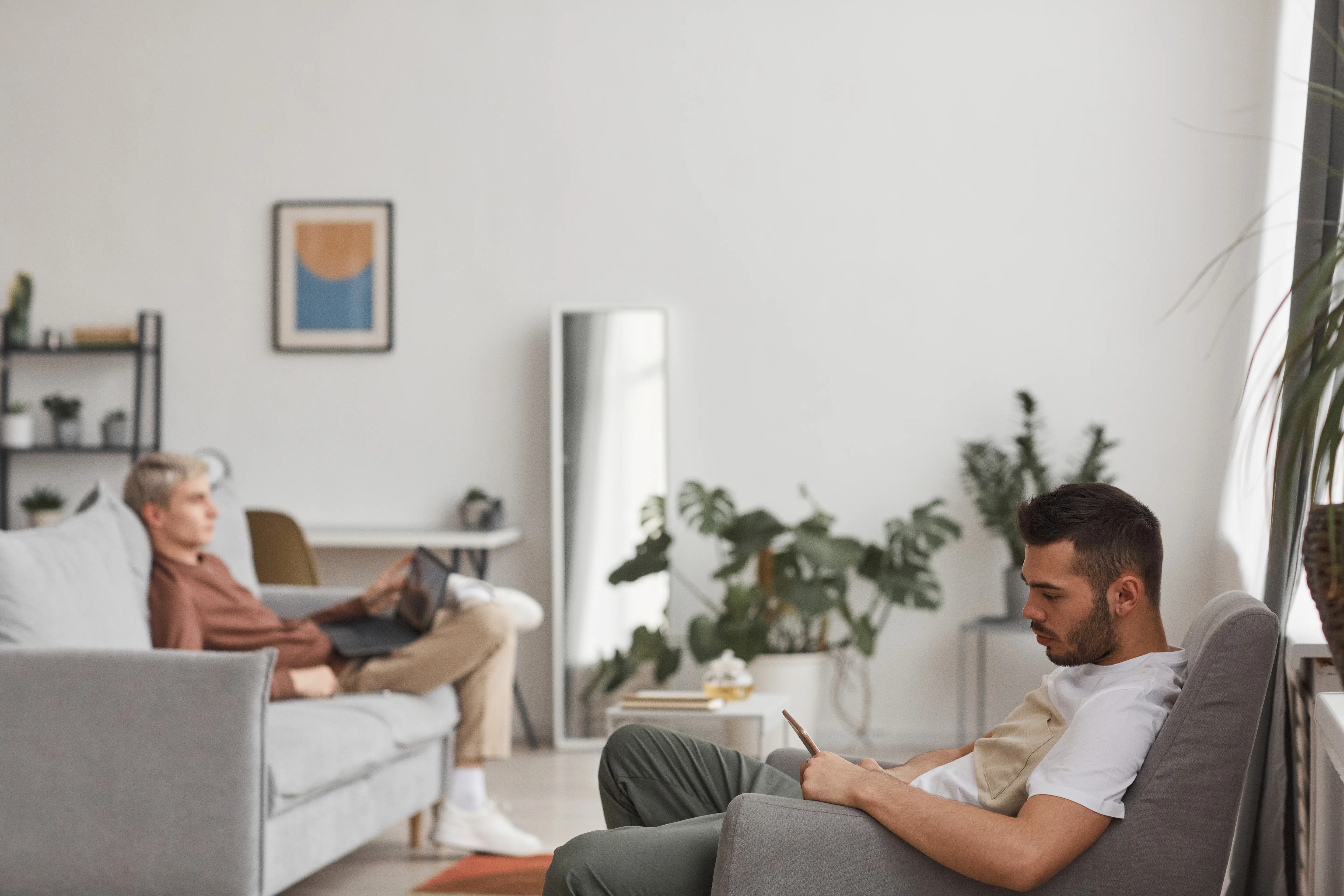 Weitwinklige Seitenansicht von zwei jungen Männern, die Internetgeräte verwenden, in einem minimalistischen Wohninterieur, mit Platz für Text.